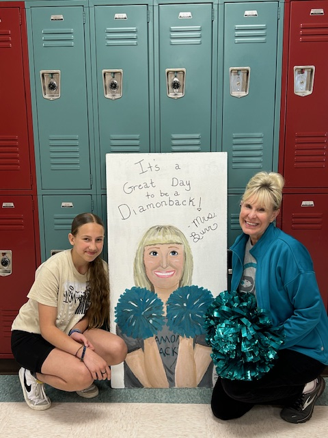 Mrs. Burr the principal with her pom poms standing next to the student who painted a  ceiling tile with Mrs. Burr's picture holding pom poms